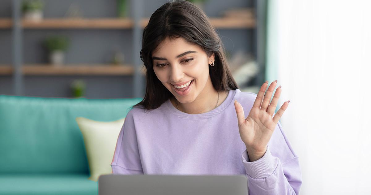 Decorativo. Una joven sonriente mira hacia abajo la pantalla de su computadora portátil y levanta la mano izquierda como si fuera a hacer una pregunta.