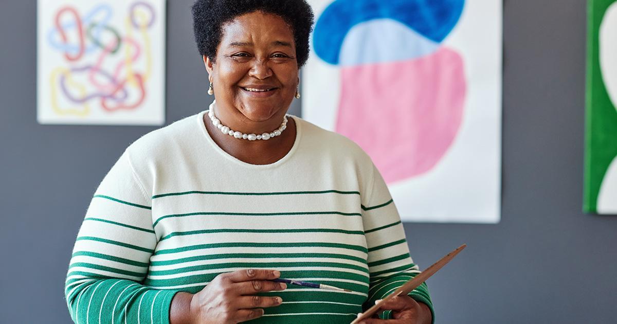 Una mujer sonriente que sostiene una paleta y un pincel de artista está frente a varios cuadros en la pared detrás de ella.