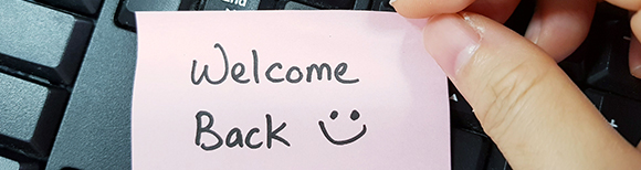 Una nota de papel en el teclado de una computadora en la que está escrito a mano, bienvenido de nuevo con una carita sonriente.