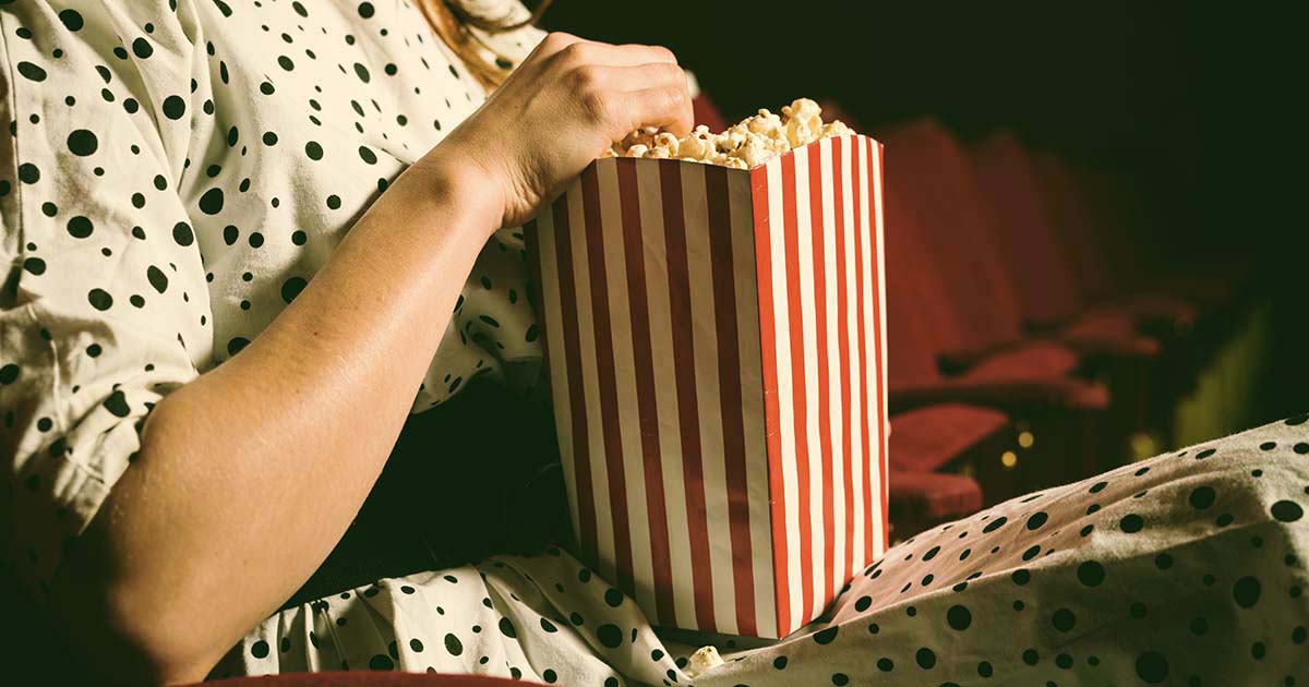 Decorativo. Una mujer que viste un vestido está sentada en un asiento dentro de un teatro oscuro. Sostiene una caja grande de palomitas de maíz en su regazo.