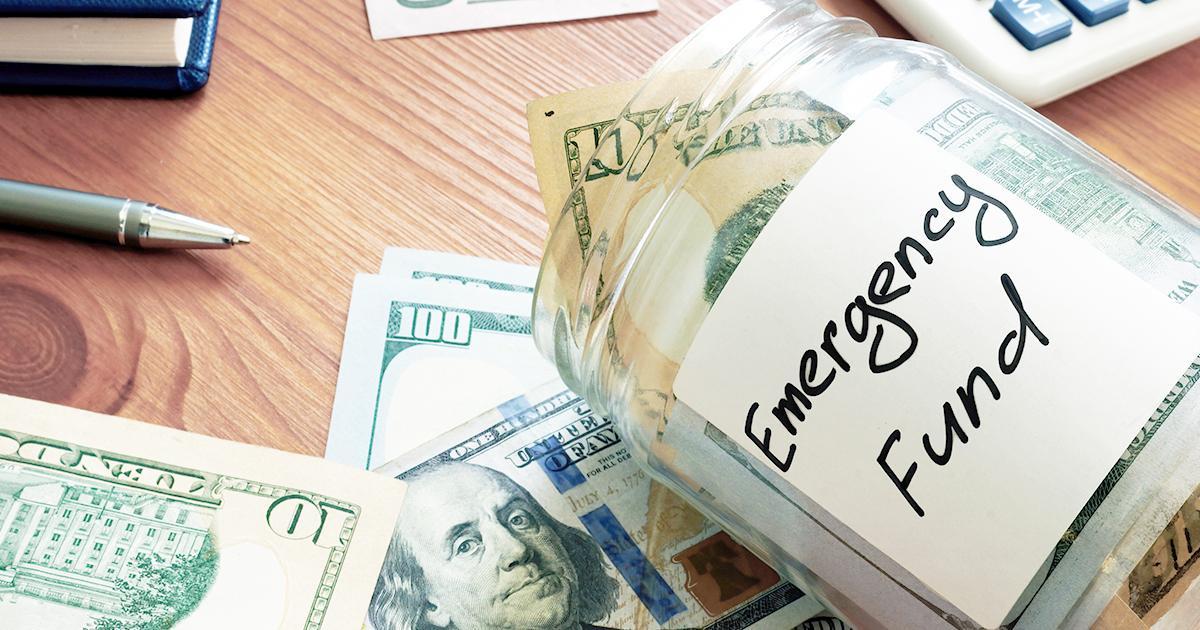 Ilustrativo. Un frasco de vidrio abierto con una etiqueta que dice “Fondo de Emergencia” se encuentra de lado sobre una mesa. Hay algo de dinero en efectivo dentro del frasco, mientras que hay más dinero en efectivo debajo del frasco. En el extremo izquierdo, hay una punta de lápiz en la esquina superior.
