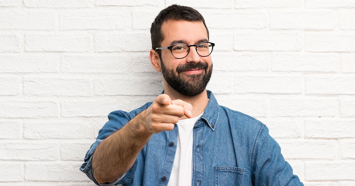 Decorativo. Un hombre con barba y anteojos sonríe y señala al espectador con el dedo índice derecho.
