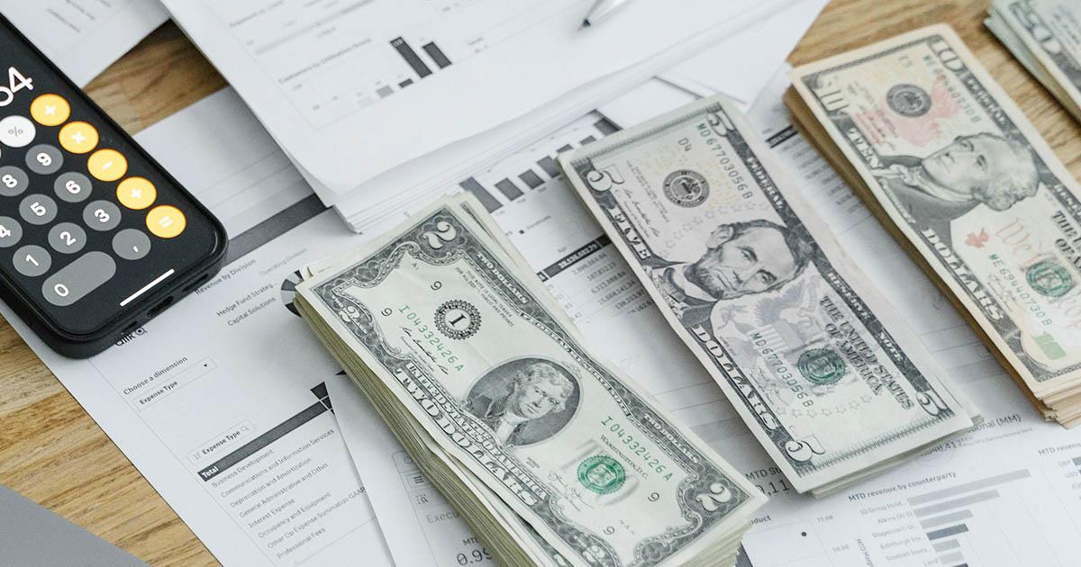 Decorativo. Una foto de tres montones de dinero en efectivo en moneda estadounidense, papeles financieros y una calculadora.