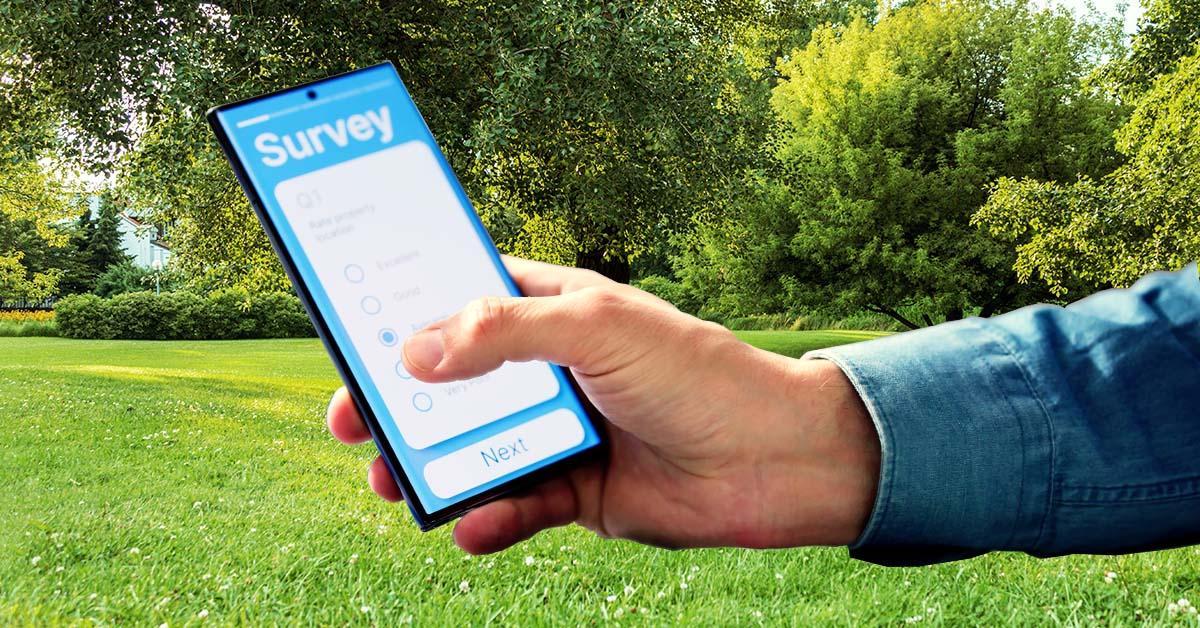 Décoratif. Une main et un poignet tenant un smartphone entrent dans le cadre par la droite. Sur l'écran du smartphone se trouve un formulaire d'enquête. En arrière-plan, une pelouse, du soleil, des haies taillées, des arbres adultes et un ciel clair avec quelques nuages.