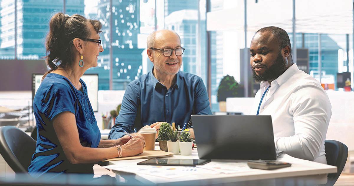 A titre d'exemple. Une femme et un homme assis dans un bureau avec de grandes fenêtres sourient à un autre homme qui regarde un écran d'ordinateur sur un bureau.