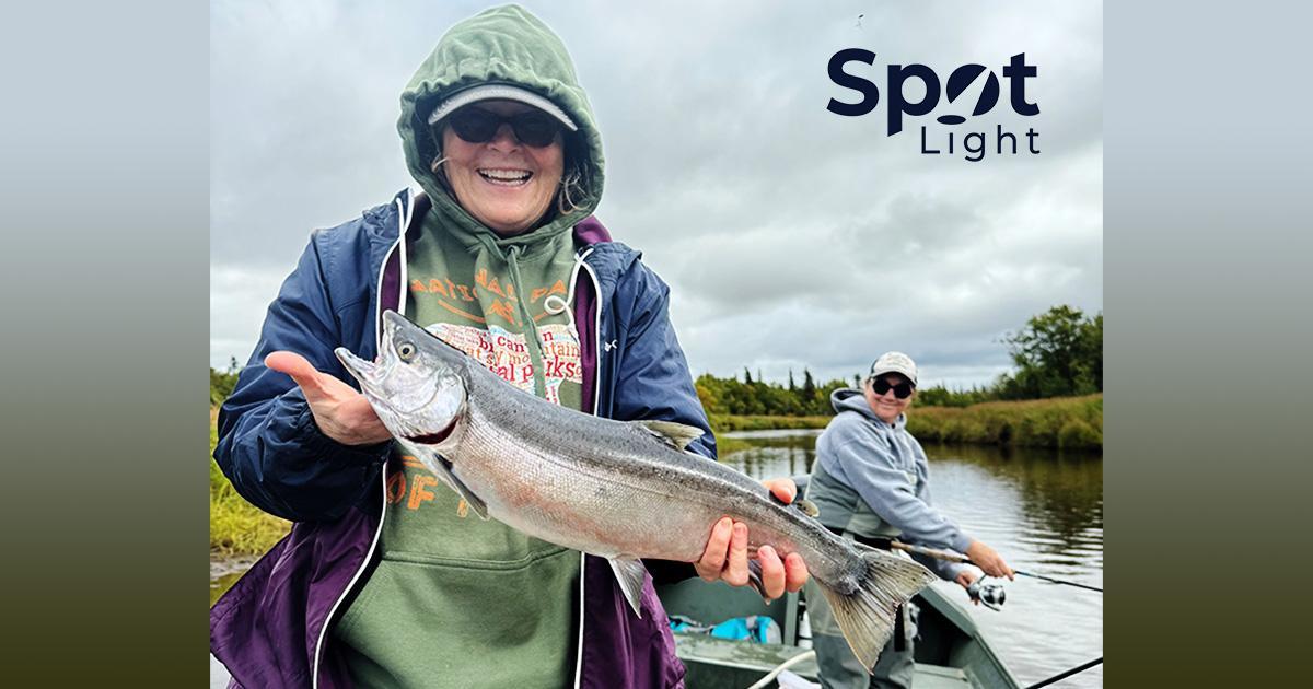 A titre d'exemple. Denise Grant se trouve dans un bateau sur l'eau et tient un saumon fraîchement pêché. Un homme à l'arrière-plan regarde. Le logo Spotlight se trouve dans le coin supérieur droit.