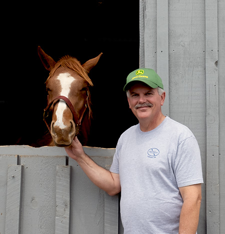 Tom Fahrney se tient à côté de son cheval Dolly's Pardon, qui regarde à travers la demi-porte de son box dans l'étable.