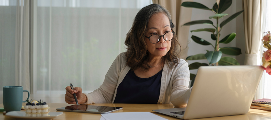 Personne assise à un bureau utilisant un ordinateur portable avec des papiers et une tablette à proximité.