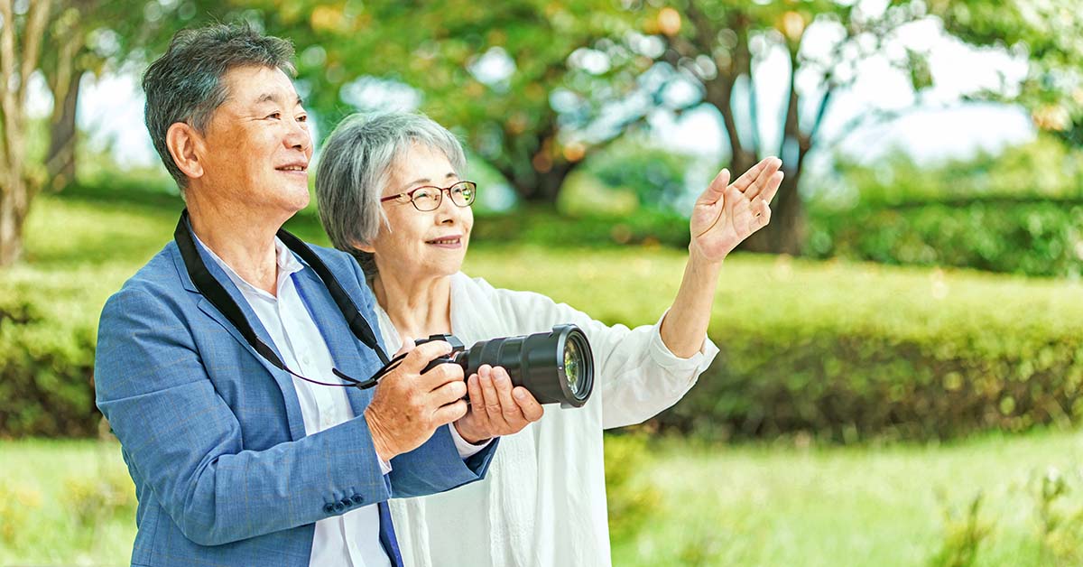 장식. 나이 든 남녀가 야외에 서 있습니다. 잘린 울타리가 배경에 있습니다. 남자는 35 밀리미터 카메라를 들고 있고, 여자는 왼손으로 프레임 밖을 향해 제스처를 취합니다.
