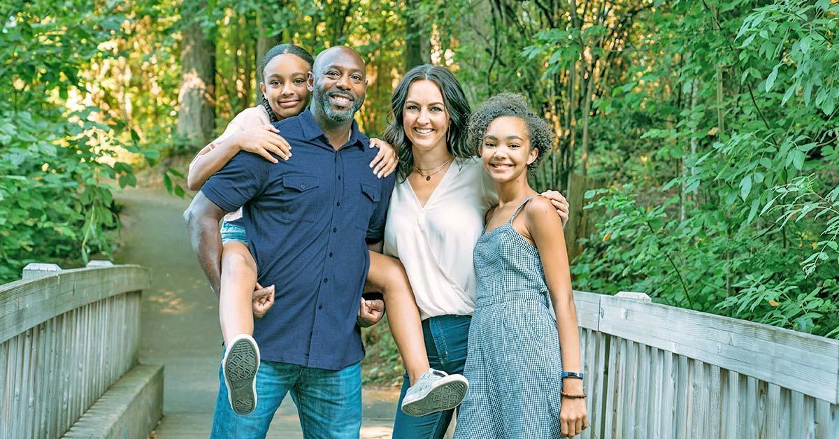 Decorative. Portrait of a family of four with a man, a woman and two young adolescent girls. They are standing on a wooden footbridge with hardwood forest on either side in the background.