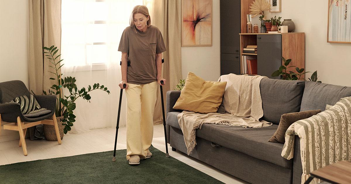 Decorative. A middle-aged woman stands supported by two canes in a residential room with a chair, large potted plant sitting on the floor, a large window with floor-length drapes, an area rug, a couch and a bookcase.