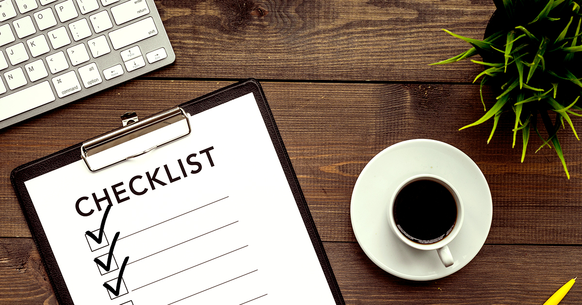 Decorative. A partial view from above of a computer tablet keyboard on a wooden surface with heavy grain texture is in the upper left corner. Below it and to the right slightly is a checklist on a clipboard with three tasks checked off. To the right of the clipboard is a cup of black coffee on a saucer. In the upper left corner is a small, spikey potted plant. Below it is a writing pen.