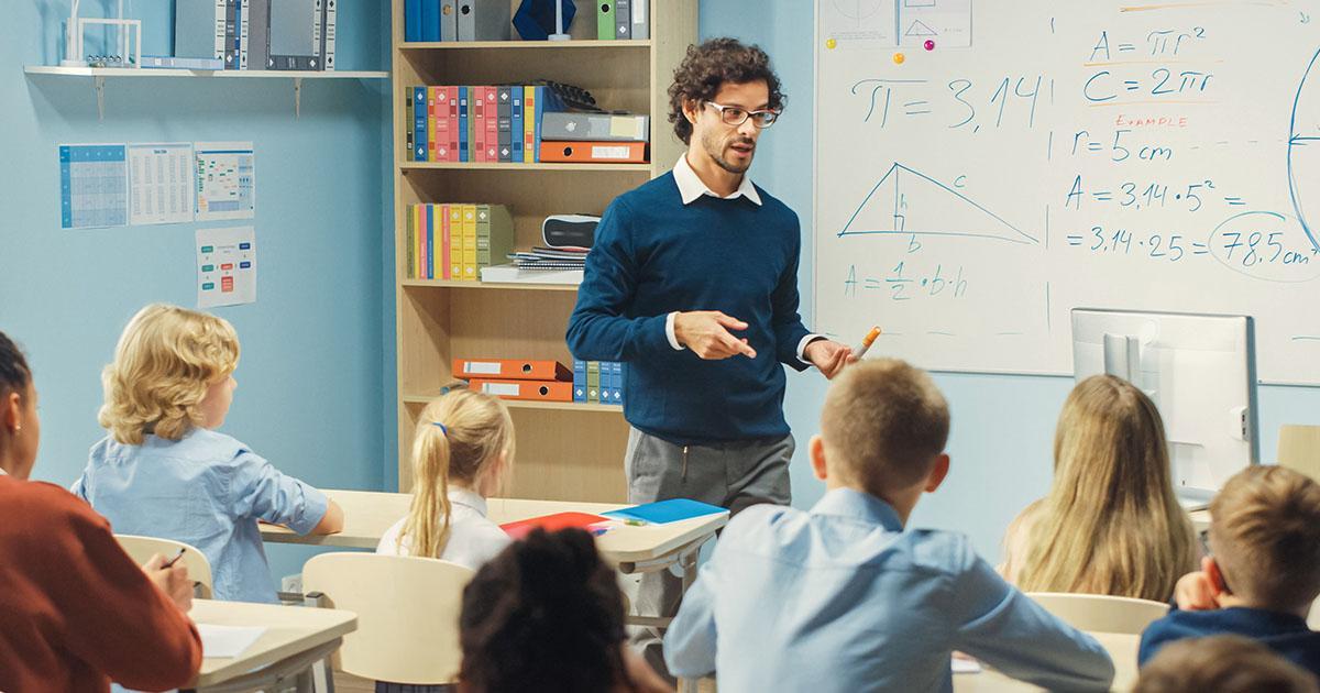 Decorative. A young teacher stands at the front of his classroom while speaking to his young students seated at desks. A whiteboard is on the wall behind the teacher.