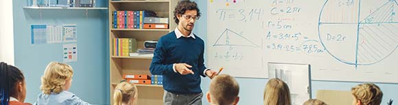 Decorative. A young teacher stands at the front of his classroom while speaking to his young students seated at desks. A whiteboard is on the wall behind the teacher.