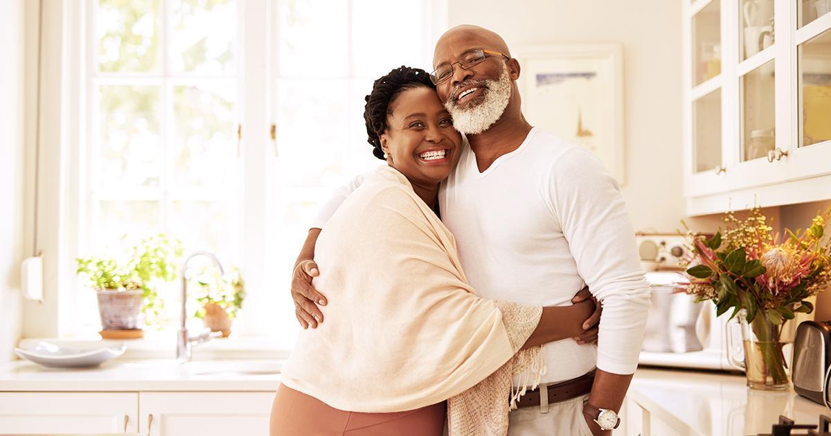 Decorative. An older couple smiles and embraces in their large, modern kitchen.