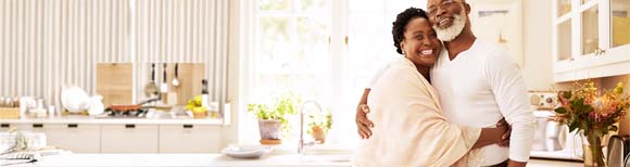 Decorative. An older couple smiles and embraces in their large, modern kitchen.