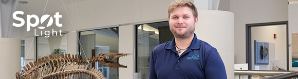 Illustrative. A photo portrait of Marshall Boyd standing on a mezzanine in the Virginia Natural History museum that overlooks a fossilized animal skeleton hanging from the ceiling.