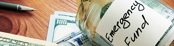 Illustrative. An open glass jar with a label reading Emergency Fund lies on its side on a tabletop. Some cash is inside the jar while more cash lies under the jar. On the far left, is a pencil point in the top corner.