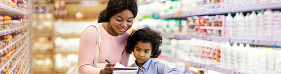 Decorative. A mother and child shop in a grocery store, pushing a grocery cart. The mother is smiling and showing the child how to use a grocery list written in a notebook.