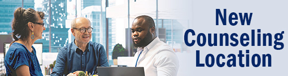 A woman and a man seated in an office with large windows smile at another man who is looking at a computer screen on a desk.