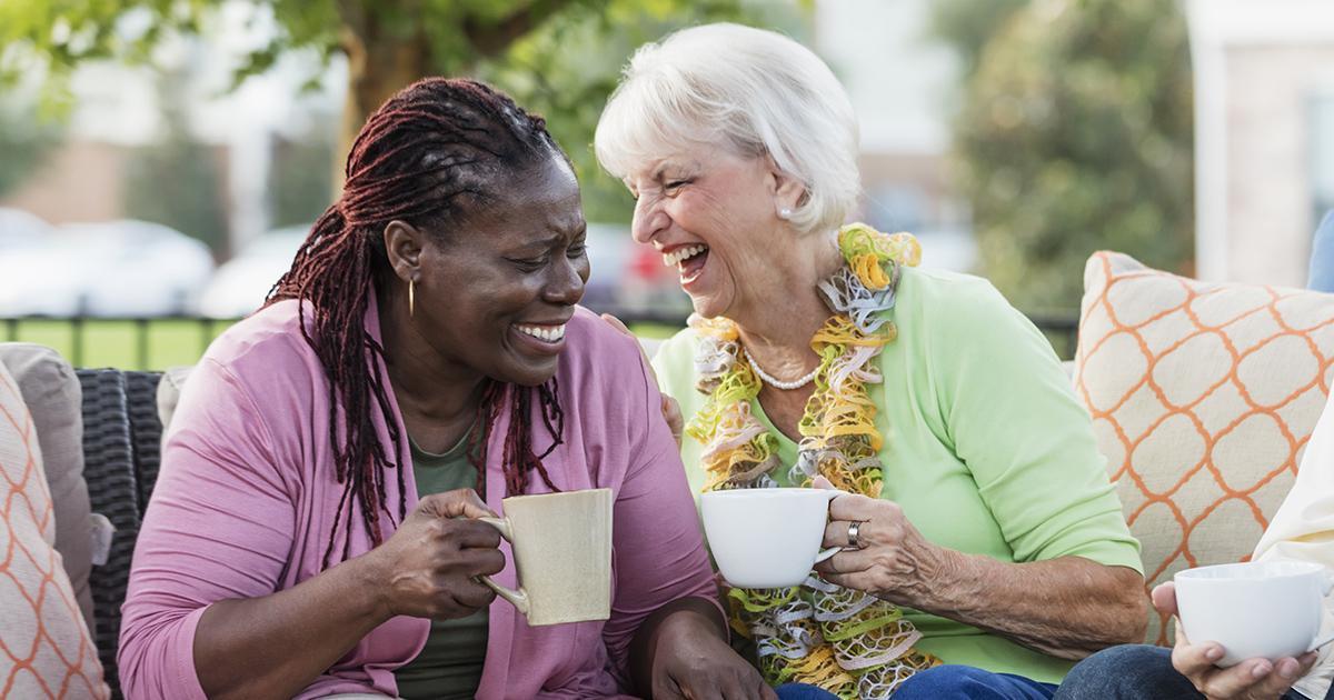 Decorative. Two older women sit on an outdoor couch with many pillows on a patio while drinking coffee and laughing.
