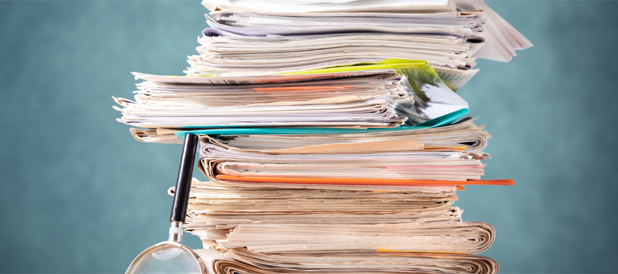 Tall stack of paper documents with folders and a magnifying glass.