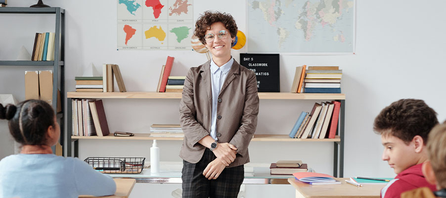 Teacher stands smiling at the front of a classroom with students seated and maps on the wall.