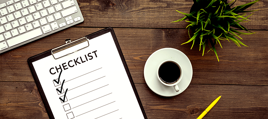 Clipboard with a checklist and marked checkboxes on a wooden desk beside a keyboard, coffee cup, plant, and pen.
