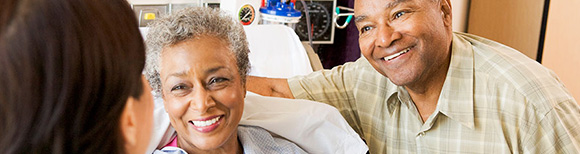 An older woman in a hospital bed and an older man beside her smile broadly at a woman in the foreground.