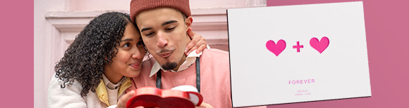 a woman sitting close to a man looks affectionately at him while he looks into a heart shaped box. Next to their photo is an envelope with hearts printed on it.