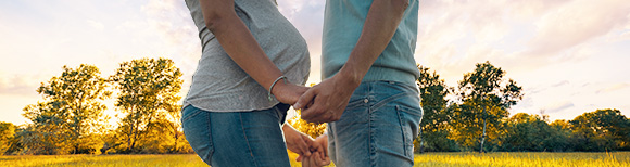 A man faces and holds the hands of a pregnant woman while the sun sets behind them.
