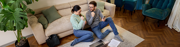 A couple wearing jeans seated on the floor in front of a couch with a laptop computer infront of them smile at each other and pump their fists in the air in celebration.