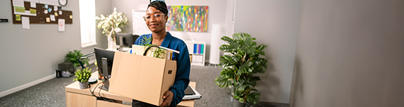 A woman smiles at the viewer while holding a box full of her belongings while she stands in a large office with several desks.