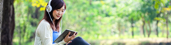 Decorative. A young woman sits on a lawn under mature trees. She is wearing headphones and looking down at a computer tablet in her hands.
