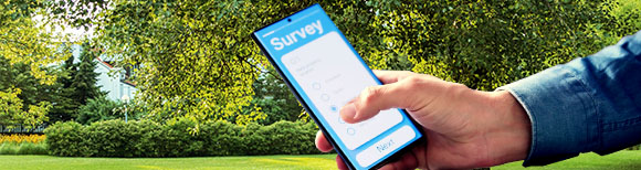 Decorative. A hand and wrist holding a smartphone enter the frame from the right. On the screen of the smartphone is a survey form. In the background is a lawn, sunshine, clipped hedges, mature trees and a clear sky with a few clouds.