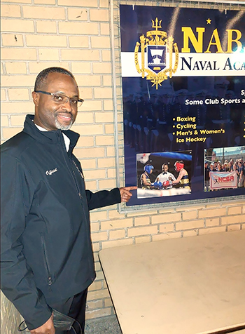 Boxing referee Brent Bovell stands next to a display case pointing to a photo of himself officiating in the ring.