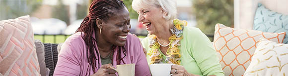 Two older women sit on an outdoor couch with many pillows on a patio while drinking coffee and laughing.