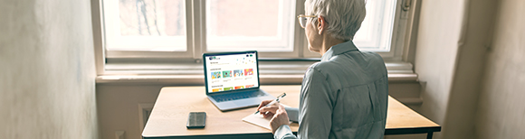 Rear view of an older woman seated at a desk taking hand written note while looking at a website on a tablet with keyboard attachment.