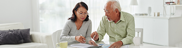 An Asian couple seated at a table in their home look over paperwork together.