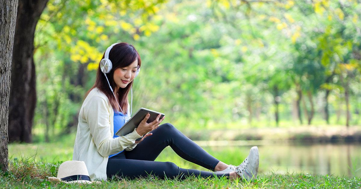 Pandekorasyon. Isang dalaga ang nakaupo sa damuhan sa ilalim ng mga puno na may sapat na gulang. Nakasuot siya ng headphone at nakatingin sa isang computer tablet sa kanyang mga kamay.