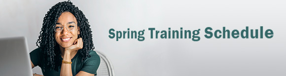 ایک مسکراتی عورت ایک میز پر بیٹھی ہے جس کے سامنے لیپ ٹاپ کمپیوٹر ہے۔ اس کے پیچھے دیوار پر موسم بہار کی تربیت کا شیڈول ٹیکسٹ ریڈنگ ہے۔