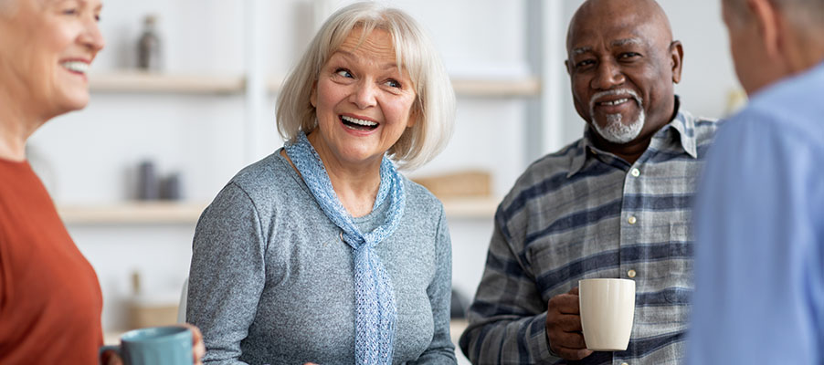四位年长者在室内手持马克杯交谈，明亮舒适的生活空间内背景有书架。